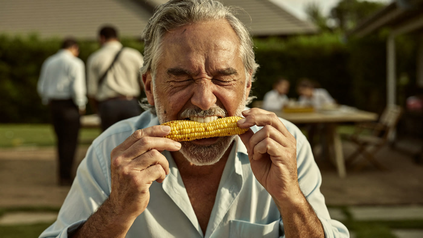 An older man happily eating corn on the cob at a family barbecue